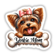 Adorable Yorkshire Terrier puppy wearing a pink bow, peeking from behind a bone-shaped banner that reads "Yorkie Mom"