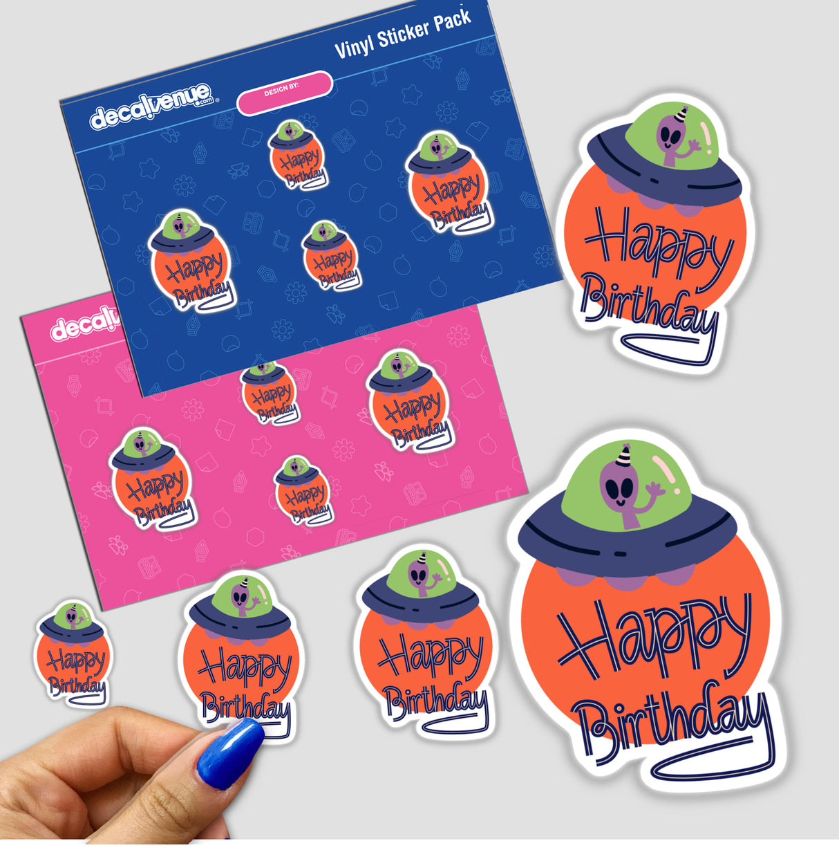 Hand holding a Happy Birthday with UFO and Alien sticker, featuring a cartoon alien in a UFO. Surrounding are similar themed cards and stickers.